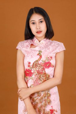 Portre kapanış stüdyosu bin yıllık pembe Çin cheongsam qipao geleneksel tavus kuşu desenli elbise içinde Asyalı kadın modelin kahverengi arka planda reklamcılık pozuyla kameraya bakın..