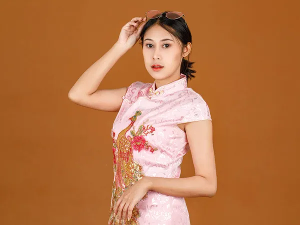 肖像画の美しさアクションミレニアルアジアの女性モデルでピンクChongsam Qipao伝統的な祭り孔雀や花のパターンドレスや眼鏡頭の上に立って笑顔ポーズ茶色の背景に — ストック写真