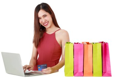 Asyalı, güzel, gülen bir kadın. Moda kıyafeti giyiyor. İnternetten alışveriş yapmak için bilgisayarını kullanıyor. Masada renkli kağıt torbaları var, izole edilmiş beyaz arka planda duruyor.