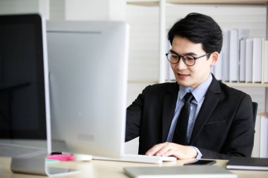 Asyalı yakışıklı, kısa saçlı, erkek, başarılı işadamı kravat, gri gömlek ve gözlüklü resmi takım elbise giyiyor. Ofis masasında çalışırken büyük ekran monitörüne bakarak gülümsüyor..