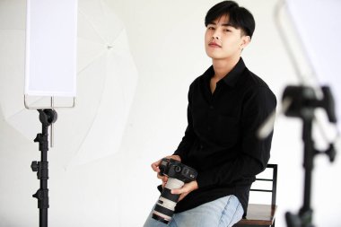 Asyalı, yakışıklı, başarılı erkek model fotoğrafçı siyah uzun kollu gömlek ve kot pantolon giyiyor. DSLR kamerayı elinde tutarken stüdyo kamerasına bakarak uzun sandalyede gülümsüyor..