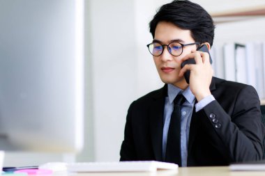 Asyalı yakışıklı iş adamı gözlüklü, kravatlı resmi siyah takım elbiseli, çalışırken, konuşurken ya da cep telefonuyla iletişim kurarken mutlu bir şekilde gülümseyen.