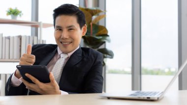 İş yerinde laptopla ve tabletle masada otururken gülümseyen ve kameraya bakan Asyalı olumlu iş adamı