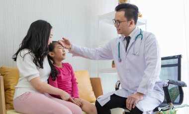 Gözlüklü genç Asyalı adam steteskopla gıdaklıyor ve annesinin yanında oturan küçük kızın alnına dokunarak muayene ediyor. Çocuk ve aile sağlık hizmeti kavramı.