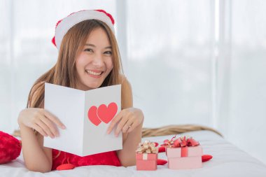 Uzun kahverengi saçlı, kırmızı ve beyaz Noel Baba şapkalı genç Asyalı kadın yatağına uzandığında kameraya gülümsüyor ve elinde küçük hediye kutularıyla mutlu bir elmas yüzük takan kırmızı bir kalp kartı tutuyor..