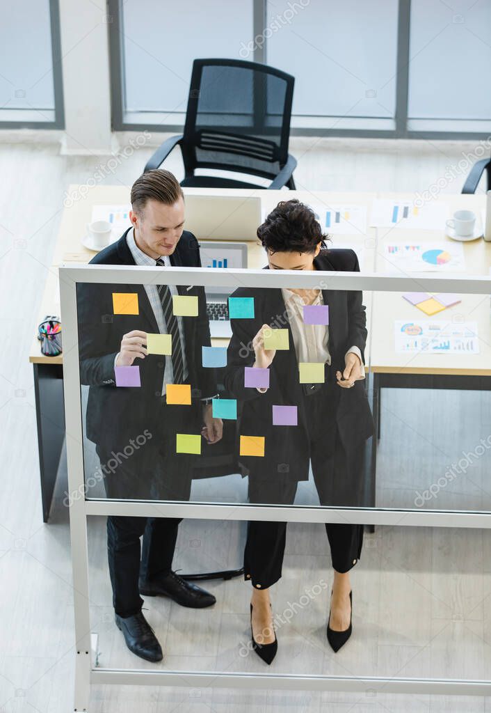 Millennial Caucasian businessman and businesswoman colleague in formal suit standing discussing consulting talking use memory colorful sticky note post-it stick on glass board in meeting room office.