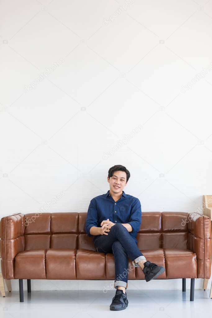 A young handsome man in blue shirt and jeans is sitting with his leg crossed at the middle on a big dark brown sofa with smile.