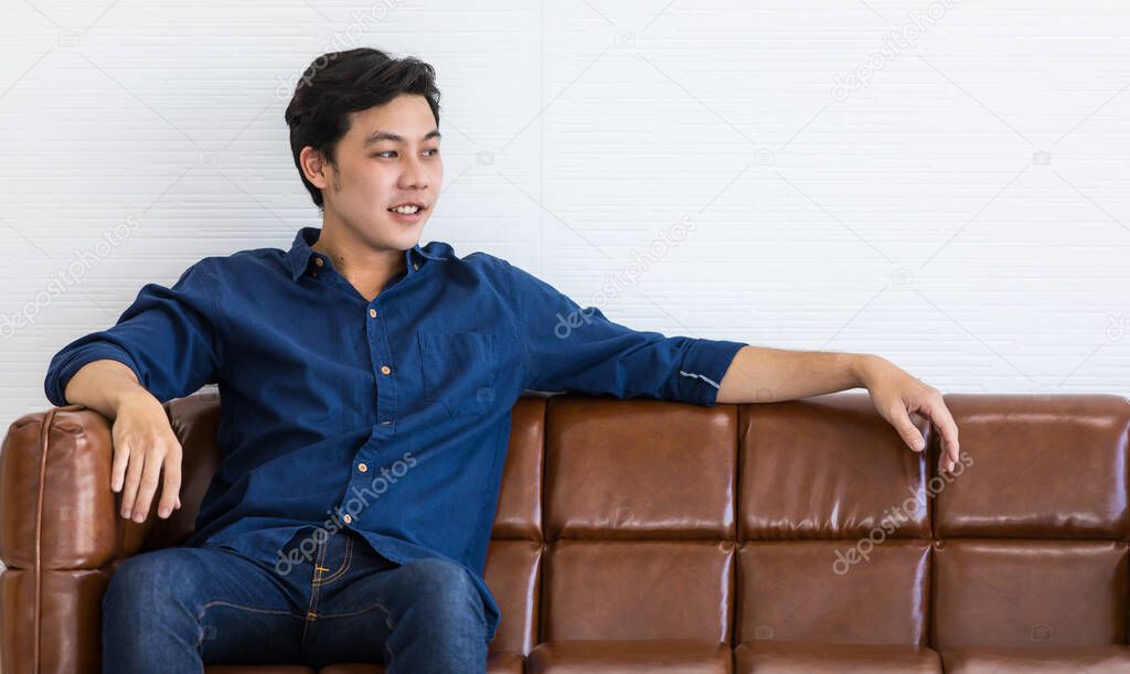 A young man in blue shirt and blue jeans is sitting on a dark brown sofa with smile. He stretch his left arm out for relax.
