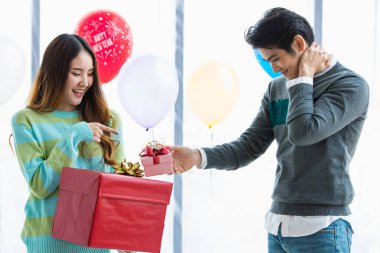 Genç Asyalı adam utangaçken elinde kırmızı bir kutu tutan, yeni yıl festivalini kutlamak için süslü balonla süslenmiş romantik bir partide gülümseyerek heyecanlanan sevimli kız arkadaşına kendinden geçmiş bir hediye veriyor.