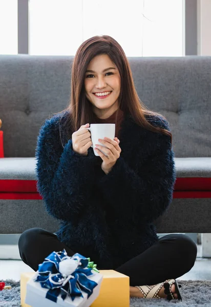 Asyalı genç bir kadın, elinde oyuncak ayılarla gri koltukta oturuyor. Elinde kahve fincanı var. Hediye kutularını kurdele ve renkli kağıtlarla süslüyor..