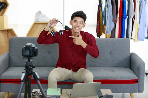 Asian Online Seller man look happy sitting at sofa holding headphones on right hand look intend and present headphones and point finger left to camera in store designed to merchant online.