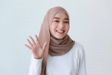 Asyalı Müslüman genç, mutlu, dindar ve tesettürlü güzel bir kadın modelin portresi. El ele tutuşup, beyaz arka planda merhaba deyin..