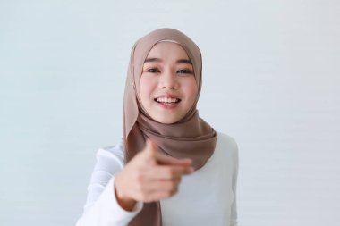 Asyalı Müslüman genç, mutlu, dindar ve tesettürlü güzel bir kadın modelin, beyaz arka planda işaret parmağıyla kameraya işaret eden tesettürünün portresi..