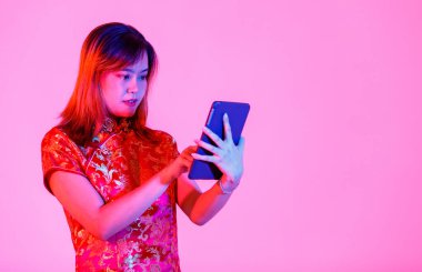 Asya 'lı genç bir genç kızın güzel kırmızı altın rengi Çin geleneksel Cheongsam Qipao tişörtü içinde beyaz arka planda mil yüzlü dokunmatik ekran tableti tutarken çekilmiş bir fotoğrafı..