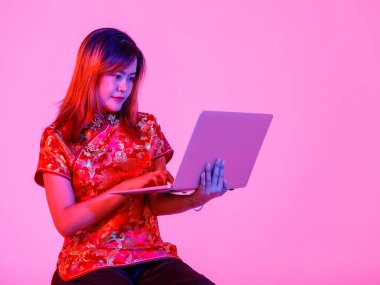 Milenyum Asyalı genç kız modelinin stüdyo fotoğrafı güzel kırmızı Çin geleneksel Cheongsam Qipao tişörtü içinde elinde dizüstü bilgisayarla pembe ışık arkaplanından alışveriş yapıyor..