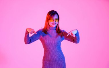 Stüdyo, gri balıkçı yaka kazaklı ve turuncu gözlüklü Asyalı genç kadın manken boş fotokopi alanını işaret ederek pembe ışık arka planında ürünü tanıtıyor..