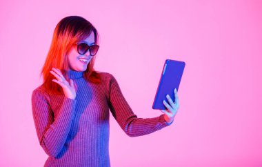Gri balıkçı yaka kazaklı, siyah gölgeli, elinde tablet bilgisayarla gülümseyen, el sallayan ve pembe ışıklı arka planda selamlaşan Asyalı genç kadın modelinin stüdyo fotoğrafı..