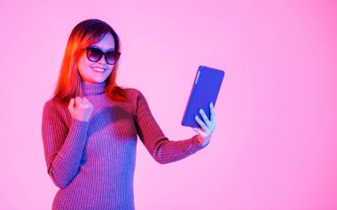 Gri balıkçı yaka kazaklı, siyah gölgeli, elinde pembe ışıklı arka planda tablet bilgisayarla gülümseyen Asyalı genç kadın modelin stüdyo fotoğrafı..