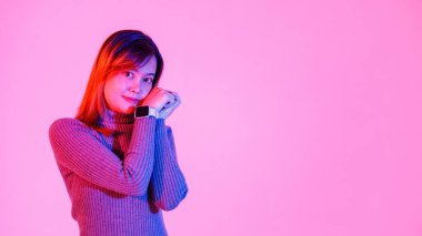 Portre stüdyosu gri balıkçı yaka kazaklı Asyalı modacı kadın mankeni çekti. Gülümsüyor, kameraya bakıyor, kolları çaprazlanmış, pembe ışıklı arka planda boş ekran akıllı saat cihazını gösteriyor..