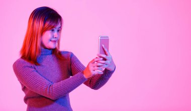 Gri balıkçı yaka kazaklı Asyalı kadın modelin portre fotoğrafı. Gülümsüyor. Dokunmatik ekranlı akıllı telefona bakıp pembe ışıklı arka planda arkadaşlarıyla sohbet ediyor..