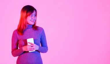 Asya 'nın modaya uygun, gri kaplumbağa yaka kazaklı, elinde dokunmatik ekranlı akıllı telefonla pembe arka plandaki boş fotokopi alanına bakarken gülümseyen kadın modelinin stüdyo fotoğrafı..