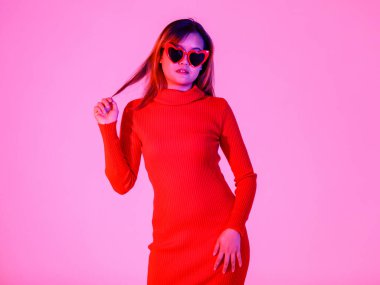 Asyalı, kendine güvenen, modaya uygun, uzun kollu balıkçı yaka kazaklı, kalp şeklinde siyah gözlüklü, beyaz arka planda seksi poz veren, Asyalı bir kadın modelin portresi..