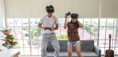 Genç ve çekici Asyalı çift ellerinde joysticklerle birlikte hoplayıp zıplamaktan ve video oyunları oynamaktan zevk alıyorlar. Oturma odasında cam pencereli eğlence ve heyecan dolu gözlükler takıyorlar..