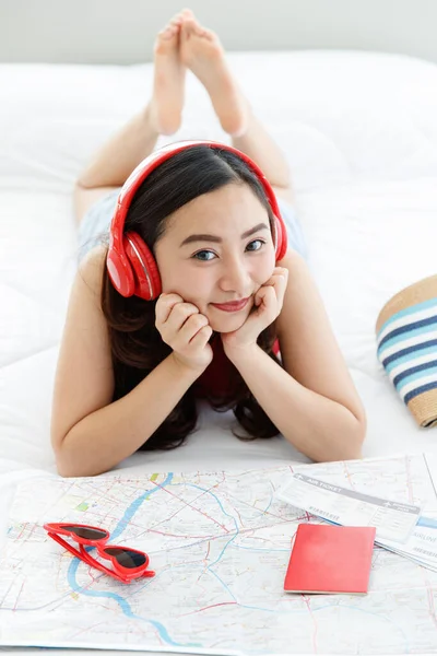 アジアの女性旅行者は大きな赤いヘッドセットを身に着けています音楽を聴く笑顔カメラでベッドの上に置くハンドバッグノートブックサングラスでベッドルーム計画休暇先紙の地図上で — ストック写真