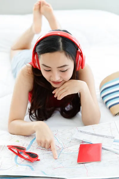 アジアの女性旅行者は大きな赤いヘッドセットを身に着けています音楽を聞いて笑顔ベッドの上に横たわりますハンドバッグノートブックサングラスで寝室の計画休暇先紙の地図上で — ストック写真
