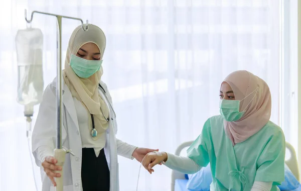 若い女性のイスラム教徒の医師は 病院の部屋で患者を回復するために使用される生理食塩水の袋のチューブを保持しながら イスラム教徒の患者の健康をチェックフェイスマスクを着用 Covid 19パンデミックの概念 — ストック写真