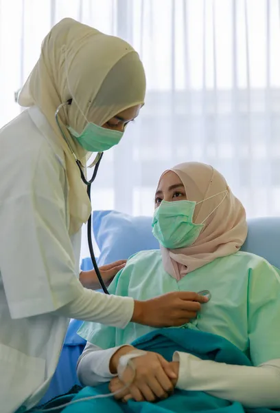 病院のベッドに座っている間 顔のマスクをした若い女性ムスリム医師が 病院の聴診器でムスリム患者の心臓をチェックします Covid 19パンデミックの概念 — ストック写真