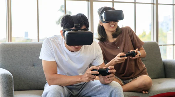 年轻迷人的亚洲夫妇手持操纵杆 喜欢一起跳着玩电子游戏 戴着Vr眼镜 带着欢乐和兴奋的心情坐在有玻璃窗背景的客厅里 — 图库照片