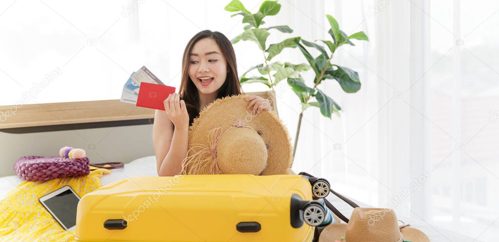 Asian young happy female traveler sitting on bed holding passport with air tickets with happy and excited actions while packing personal traveling stuff to yellow trolley baggage on bed.