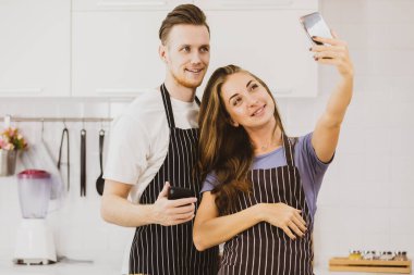 Önlüklü mutlu çift hafif mutfakta birlikte yemek pişirirken masanın yanındaki akıllı telefondan kendi portresini çekiyor.