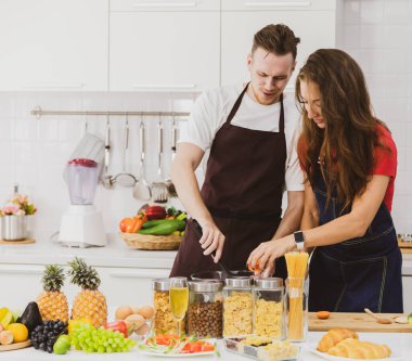 Önlüklü mutlu bir çift, çeşitli ürünlerle aynı masada otururken aynı anda açık mutfakta, modern mutfak gereçleriyle birlikte...