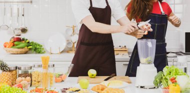 Önlüklü, tanınmayan bir çift, mutfakta çeşitli ürünlerle yemek pişirirken modern blender 'a taze malzemeler ekiyor.