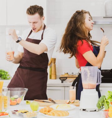 Önlüklü, çeşitli malzemelerle masada bardaklarla dans eden ve hafif modern mutfakta meyve püresi yapan pozitif bir çift.