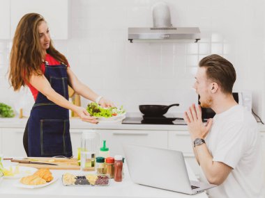Önlüklü kız arkadaşın mutfak ürünleriyle masanın yanında duran erkek arkadaşına taze salata göstermesi.