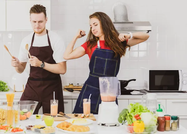 Önlüklü, çeşitli malzemelerle masada bardaklarla dans eden ve hafif modern mutfakta meyve püresi yapan pozitif bir çift.