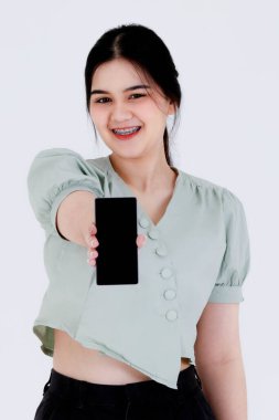Asyalı genç bayan modelin portre çekimi at kuyruğu saç stili diş teli takıyor ve beyaz arka planda boş ekranlı akıllı telefonu işaret eden en iyi kıyafetini giyiyor..