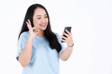 Asya tombul tombul, uzun siyah saçlı, mavi tişörtlü ve kol saatli, gülümseyen, akıllı telefon sallayan beyaz arka plandaki video çağrısında merhaba diyen kadın modelin stüdyo fotoğrafı..