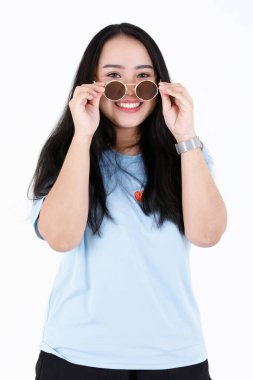 Asyalı tombul, şişman, sağlıklı, mavi gömlekli, uzun siyah saçlı, gülümseyen bir kadın modelin portresi. Elinde gülümseyerek kameraya bak. Beyaz arka planda klasik güneş gözlüklerini çıkar..