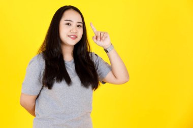 Asyalı genç tombul tombul, mutlu, uzun siyah saçlı, gri tişörtlü kadın modelin portre çekimi. Sarı arka plandaki boş fotokopi alanında gülümsüyor..