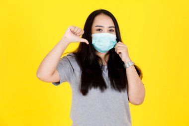 Asyalı genç tombul tombul, uzun siyah saçlı, gri giysili ve yüz maskeli bir kadın modelin portre çekimi. Yüzünde el ele tutuşurken şok olmuş sarı arka planda şaşkınlığa uğradım..