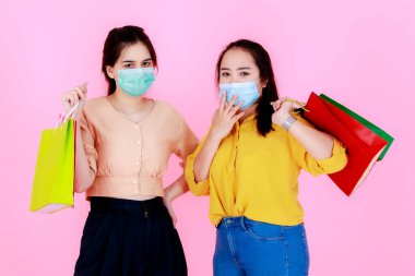 Portre stüdyosunda, pembe arka planda salgın hastalık karantinası sırasında renkli alışveriş torbaları taşıyan, yüz maskesi takan ve günlük kıyafet giyen iki Asyalı genç bayanın portresi var..