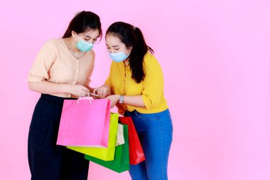 Portre stüdyosunda, pembe arka planda salgın hastalık karantinası sırasında renkli alışveriş torbaları taşıyan, yüz maskesi takan ve günlük kıyafet giyen iki Asyalı genç bayanın portresi var..