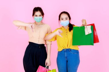 İki mutsuz Asyalı genç mutsuz bayan müşterinin portre çekimi. Yüz maskesi takan ve günlük kıyafet giyen, pembe arka planda başparmaklarını gösteren renkli alışveriş çantaları taşıyan mutsuz bayan arkadaşlar..