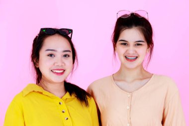 İki Asyalı, neşeli, at kuyruklu, güneş gözlüklü, güneş gözlüklü kız ya da kız kardeşlerinin pembe arka planda kameraya gülümseyişlerinin portresi..