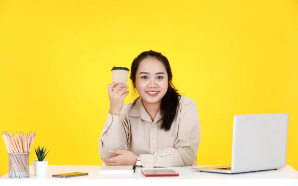Portrait studio shot of Asian chubby plump female secretary employee worker sitting take break holding paper disposable hot coffee cup in hand look at camera smiling at company on yellow background.