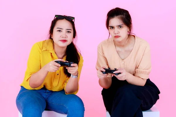 ポートレートスタジオショットの二つのアジアの若い陽気な競争力のある女性の友人や姉妹ゲーマーでカジュアルウェアでサングラス座って笑顔保持ジョイスティックゲーム一緒にピンクの背景に — ストック写真